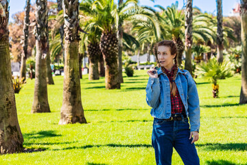 A woman in a spring denim outfit is biting shackle of glasses while standing in a spacious southern park among palm trees.