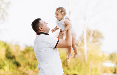 Obraz premium Father's day. Dad is hugging his baby daughter outdoors and having fun by the pond. Family enjoying life together on nature. Happy young family day. The concept of family summer holiday.