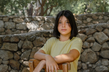 A young teenager girl sits on a wooden chair against a stone wall.