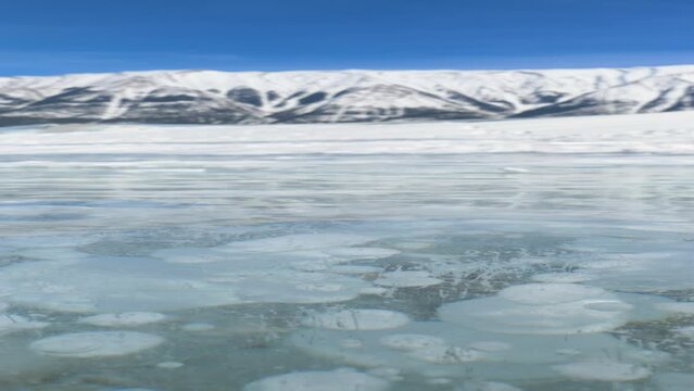 VERTICAL, CLOSE UP, DOF: Strong gusts of wind blow across the scenic frozen Lake Abraham on a sunny winter day. Stunning shot of Canadian Rockies behind a glassy frozen lake full of methane bubbles.