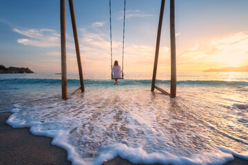 Happy young woman on wooden swing in water, beautiful blue sea with waves, sandy beach, golden sky at sunset. Summer holiday in Oludeniz, Turkey. Girl ride on a swing on sea coast, clear water. Travel