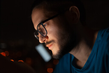 Night portrait of serious man looking in smartphone on background of window.