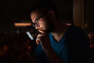 Night portrait of serious man looking in smartphone on background of window.