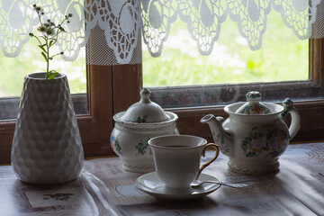 a tea set and a vase with daisies on the table in front of a wooden window of a rural house