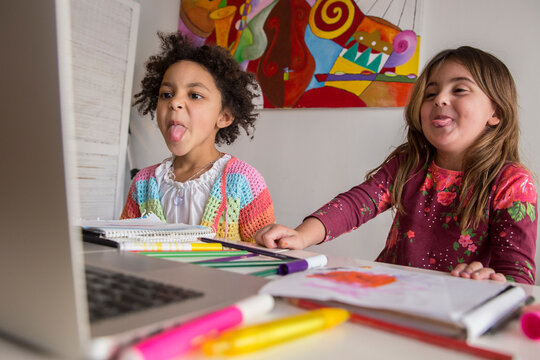 Two multiethnic little girls learning on line and sticking out tongue to the computer at home. Back to school concept.