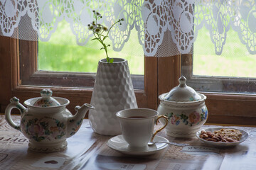 a tea set and a vase with daisies on the table in front of a wooden window of a rural house