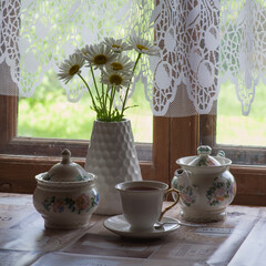a tea set and a vase with daisies on the table in front of a wooden window of a rural house