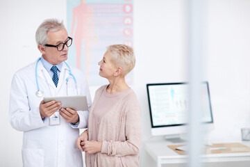 High-qualified senior gray-haired doctor in eyeglasses and lab coat standing in clinic room and counseling mature female patient