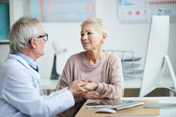Obraz premium Senior doctor in eyeglasses sitting at table and holding hand of mature female patient while giving hope and comfort to her