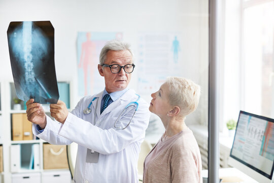 Frowning handsome aged doctor in eyeglasses holding x-ray image against light while explaining spinal problems to senior female patient