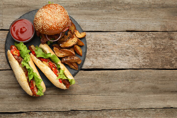 Tasty burger, hot dogs, potato wedges and sauce on wooden table, top view with space for text. Fast...