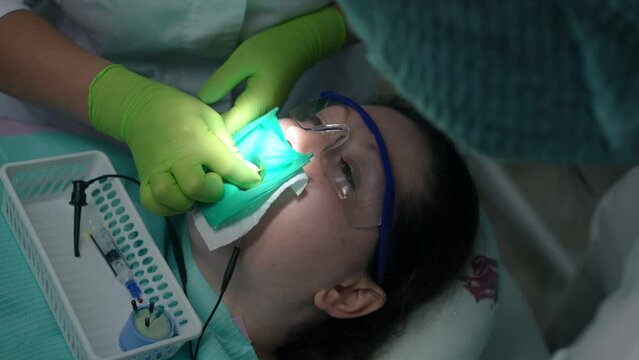 Headshot of Caucasian female patient in protective eyeglasses and dental dam with doctor and assistant treating bad tooth in slow motion. Close-up portrait of ill woman undergoing treatment in clinic