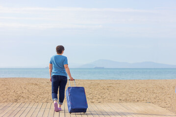 Fototapeta premium one woman in casual clothes and with a suitcase walked along the beach to the sea, back view with copy space