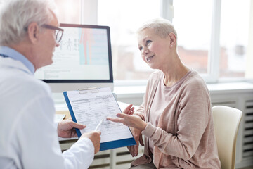 Obraz premium Smiling senior female patient with blond hair sitting at table and pointing at paper in clipboard while asking doctor about prescribed drugs