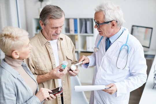 Serious Aged Male Doctor In Glasses Standing In Clinic And Explaining Senior Couple How To Take Medicine Correctly