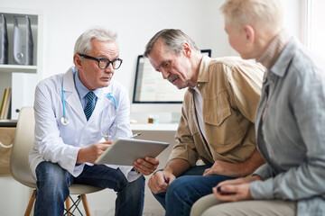 Serious senior male doctor in white coat using digital tablet while analyzing tests results of aged...