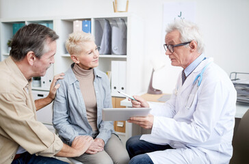 Serious high-qualified cancer doctor in glasses showing medical records on tablet and giving advice to mature woman with serious disease, her husband holding her hand as symbol of support