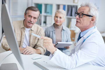 Plakat Confident qualified gray-haired doctor in white coat sitting at table and using modern computer while showing results of medical checkups to senior patient