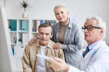 Mature man embraced by wife sitting in doctors office and listening to doctors explanation while getting good medical tests results at appointment
