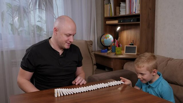 A little blond boy with his father starts a row of dominoes on a wooden table, the domino bones fall.