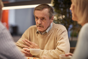 Wrinkled aged man in beige sweater sitting at table and drinking tea while sharing stories with friends