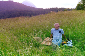 Fototapeta na wymiar mature man, senior sits on gentle slope of mountain, next to picnic basket with fruit, thermos, green grass in meadow, concept of family picnic on nature, enjoy life in old age, active lifestyle