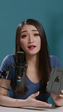 Vertical video: POV of asian woman doing product review with v mount battery in studio, filming portable v lock accumulator recommendation on vlogging camera. Rechargeable power plug on vlog. Tripod