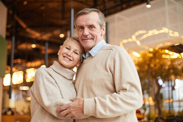 Portrait of happy married elderly couple in casual sweaters embracing each other in modern...