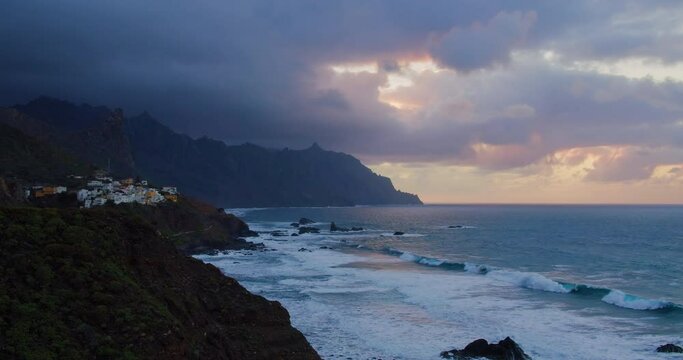 Silhouette cliffs with buildings of little tropical town Almaciga in sea revealing sunset. Ocean waves roll beautifully on a rocky shore. Tenerife.