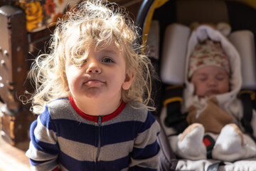 Closeup selective focus shot of cheeky two year old caucasian boy with wavy blonde hair and blurry newborn sister in baby seat in background.