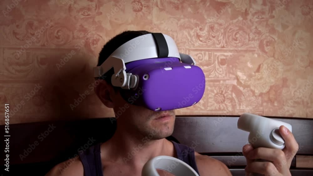 Poster man in a virtual reality helmet controls the controllers. a young man at home sitting on a bed is in