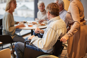 Mature man in wheelchair pushed by wife joining friends in cafe, group of seniors drinking tea in background