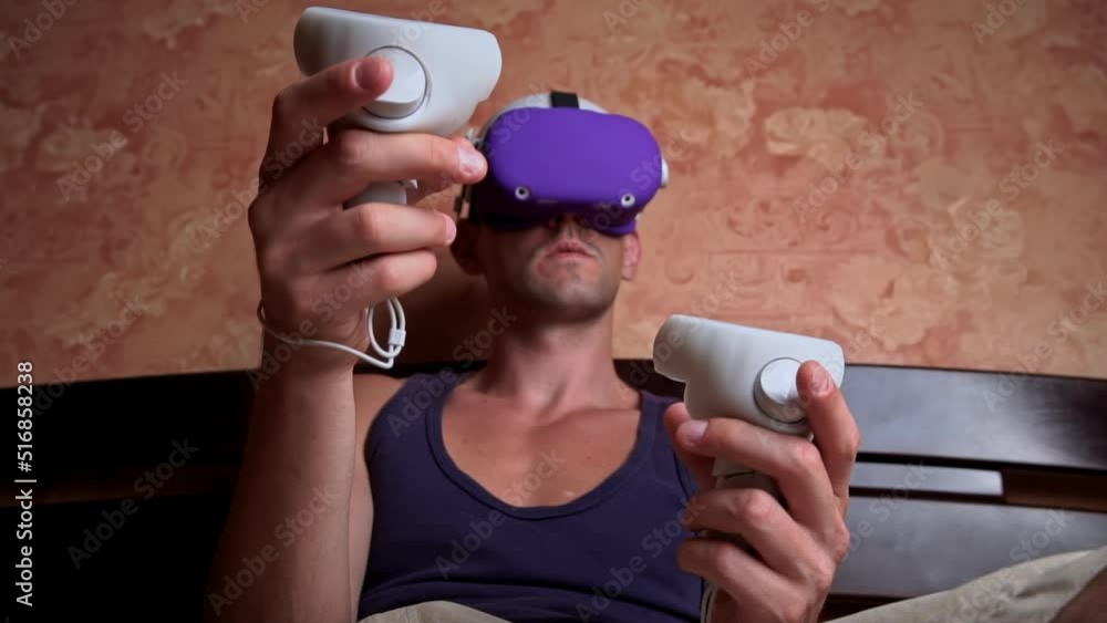 Poster man in a virtual reality helmet controls the controllers. a young man at home sitting on a bed is in