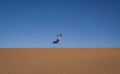 Naklejka na ściany i meble Man jumps over the sand dunes. Colorful wide shot of a funny moment in a desertic landscape 