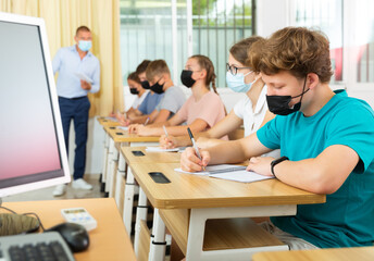 Pupils in face masks sitting in class and listening carefully to male teacher.