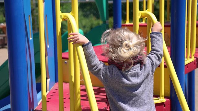 Camera panning around the rear of a two year old boy with blonde wavy hair playing at a recreational park, climbing the steps to a slippery slide.