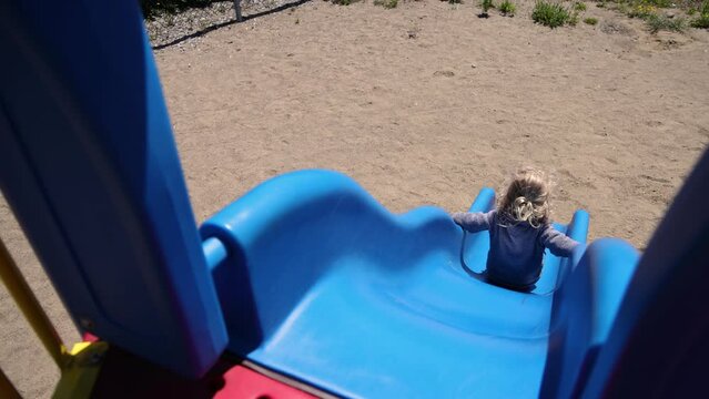 Slow motion view from top of a blue playground slide as a happy three year old boy sits and slides down to sandy ground at bottom. With copy space.
