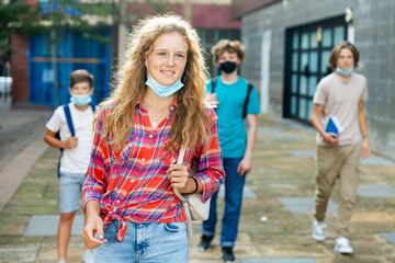 Teenager girl in face mask going home after classes in school with other teens who walking in background. .