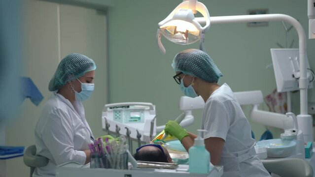 Side view positive dental assistant using curing light for filling polymerization talking with dentist in slow motion. Medium shot of Caucasian nurse and doctor curing tooth of patient indoors