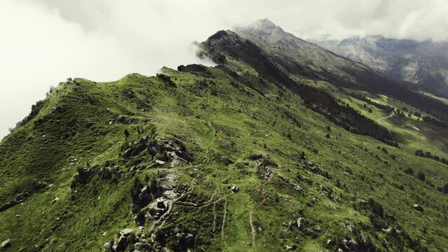 Aerial drone shot of a grass covered mountain top with a small trail leading along. More green mountain ranges in the background with clouds around the peaks on a sunny day. 4K flying forward.