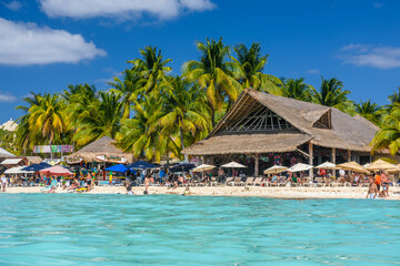 People sunbathing on the white sand beach with umbrellas, bungalow bar and cocos palms, turquoise...
