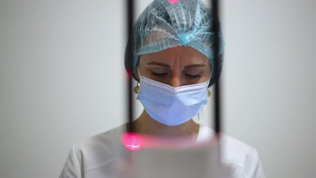 Concentrated Caucasian woman in face mask thinking analyzing X-ray image of teeth and jaw standing at cone beam scanner. Front view portrait of professional focused dentist radiologist in hospital