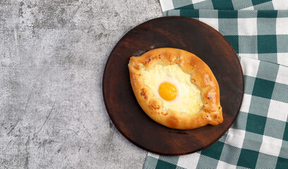 Caucasian flat bread - Adjarian khachapuri with cheese and egg on a round wooden cutting board on a dark grey background. Top view, flat lay.