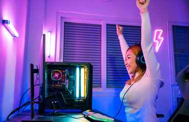 Winning Victory. Asian gamer playing online video game excited on desktop computer PC colorful neon LED lights, young woman in gaming headphones using computer she happy successful, E-Sport concept - obrazy, fototapety, plakaty