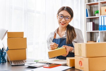 Looking Camera, Startup small business entrepreneur or freelance Asian woman using a laptop with box, online marketing packaging box and delivery, SME concept.