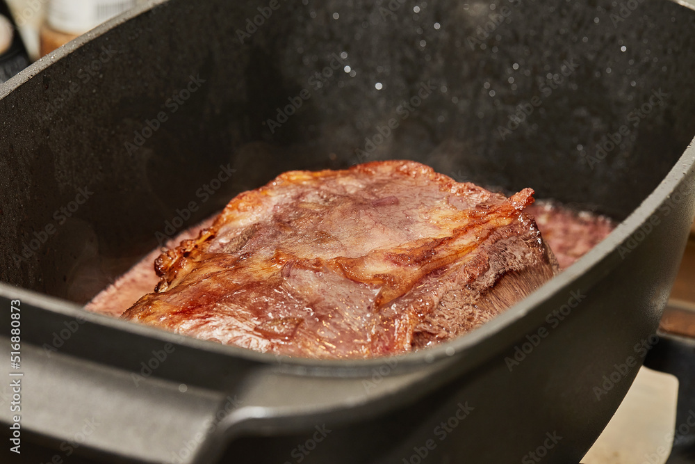 Wall mural Piece of meat is fried with onions in wine sauce in rectangular pan over fire on gas stove