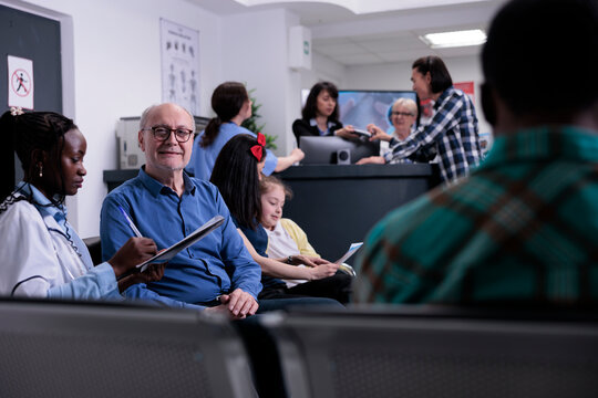 Portrait Of Older Man With African American Medical Doctor Looking At Patient Data While Diverse People Are Talking At Hospital Reception. Senior Citizen In Busy Hospital Clinic Attending Appointment.