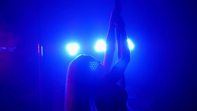 A flexible dancer in lingerie dances on a pole under the light of multi-colored spotlights in a night dance club.