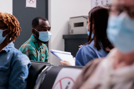 Male Patient With Face Mask Talking To Assistant, Sitting In Waiting Area At Hospital Reception Lobby. Man Doing Healthcare Consultation With Medical Worker, Nurse And Person At Facility.