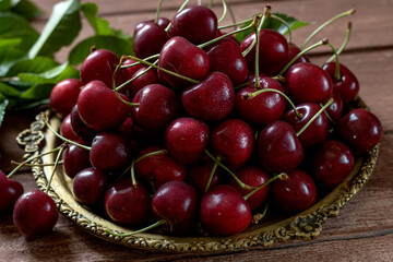 A metal vintage dish filled with cherries, cherry leaves nearby.